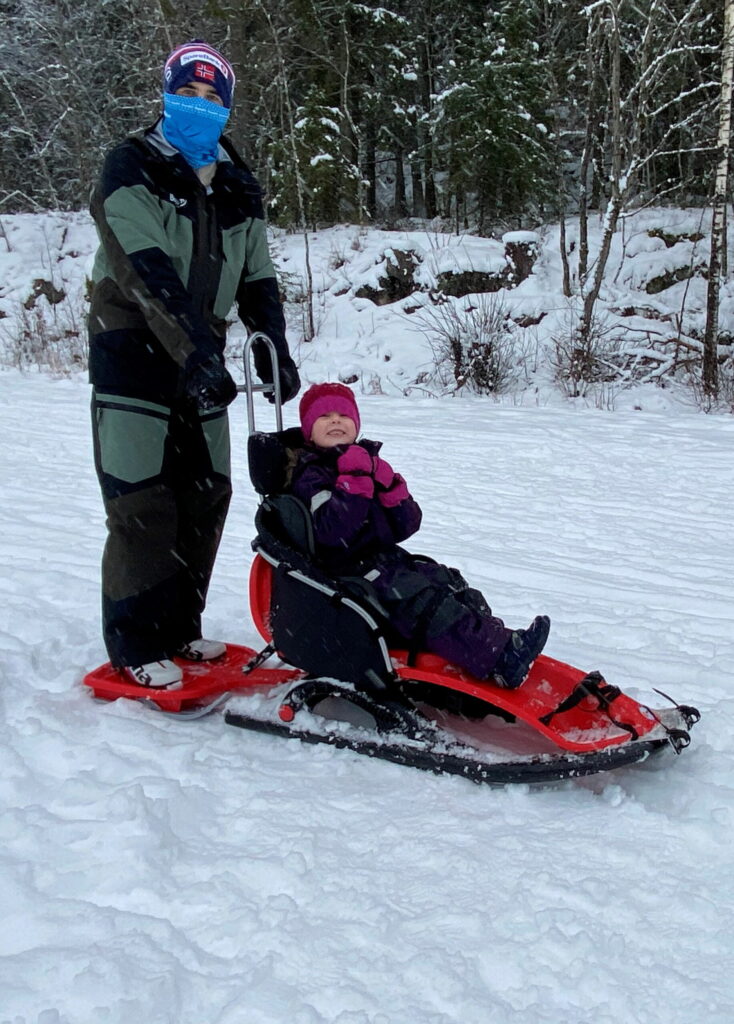 Mads og Oline er klare for aketur med SnowComfort. Det snør lett, Mads står på ståbrettet bak Oline. Oline har armene opp under ansiktet og ser spent ut.