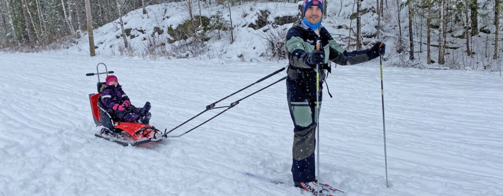 Mads og Oline på skitur i snøvær. Mads står foran og drar SnoComforten/pulken med Oline sittende i