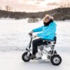 En dame med blomst i håret ser på kameraet. Hun sitter i en Eloflex s1 scooter. Det er vinter, og bakken er dekt med snø. Damen har på seg godt med klær.