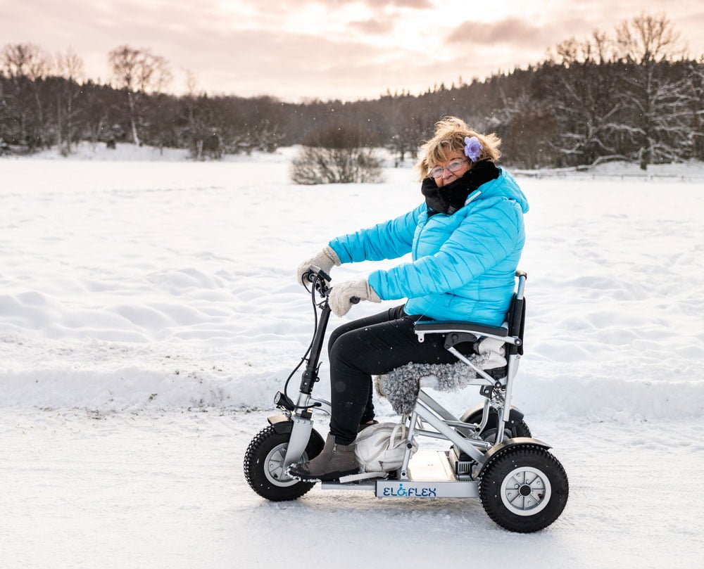 En dame med blomst i håret ser på kameraet. Hun sitter i en Eloflex s1 scooter. Det er vinter, og bakken er dekt med snø. Damen har på seg godt med klær.