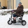 En dame sitter i scooteren Eloflex S1. Bildet er tatt fra siden. Damen har på seg vinterklær. Bildet er tatt utenfor en blokk. Det er snø på bakken og på hekken.
