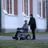Et eldre ektepar på tur. Den eldre damen sitter i en Eloflex elektrisk rullestol. Mannen holder i kjørehåndtakene bakpå. De står utenfor et stort bygg.