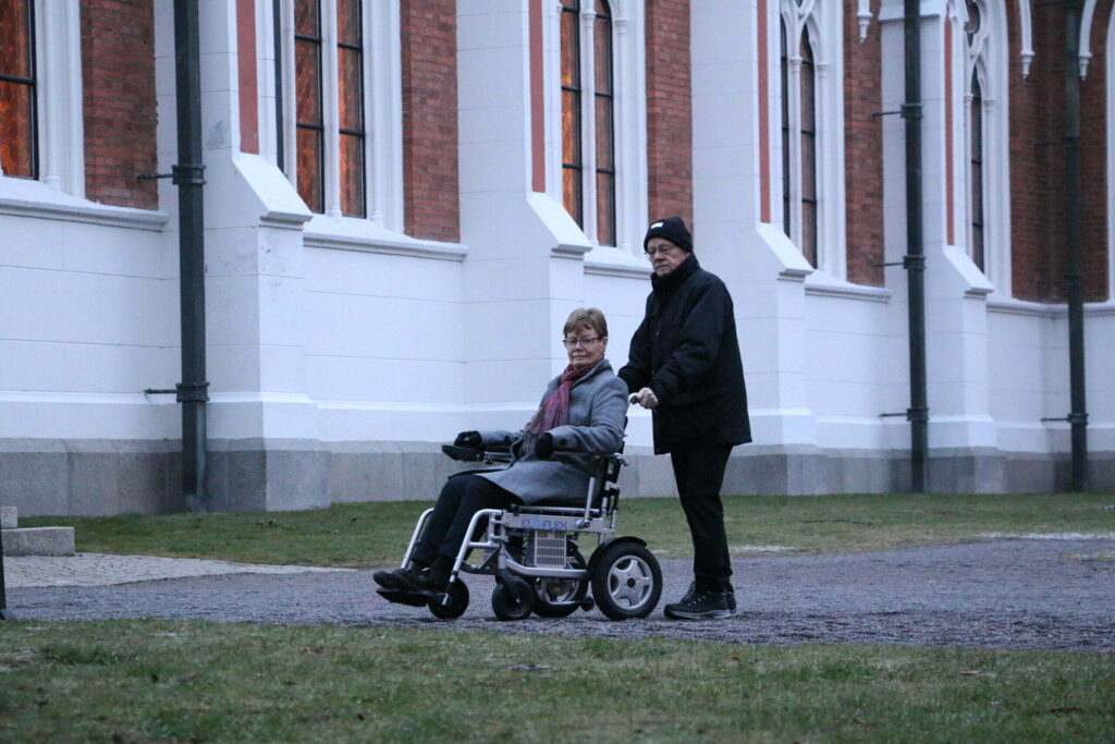 Et eldre ektepar på tur. Den eldre damen sitter i en Eloflex elektrisk rullestol. Mannen holder i kjørehåndtakene bakpå. De står utenfor et stort bygg.
