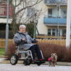 Eldre dame kjører sin Eloflex X elektriske rullestol i nabolaget sitt. Hun går tur med hunden, og i bakgrunnen ser du noen hager og en blokk.