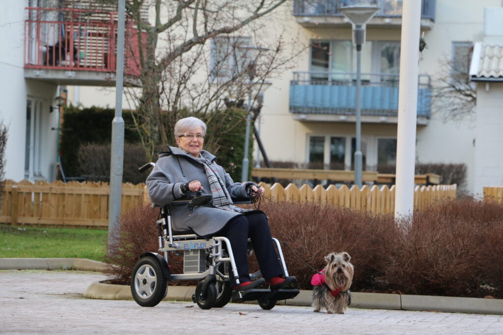 Eldre dame kjører sin Eloflex X elektriske rullestol i nabolaget sitt. Hun går tur med hunden, og i bakgrunnen ser du noen hager og en blokk.