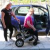 Bildet viser en dame som er på vei ut av sin Eloflex P elektriske rullestol. Hun er på vei inn i bilen sin. En annen dame holder tak i el-rullestolen på baksiden. De er kledd i bukse og t-skjorte og sola skinner.