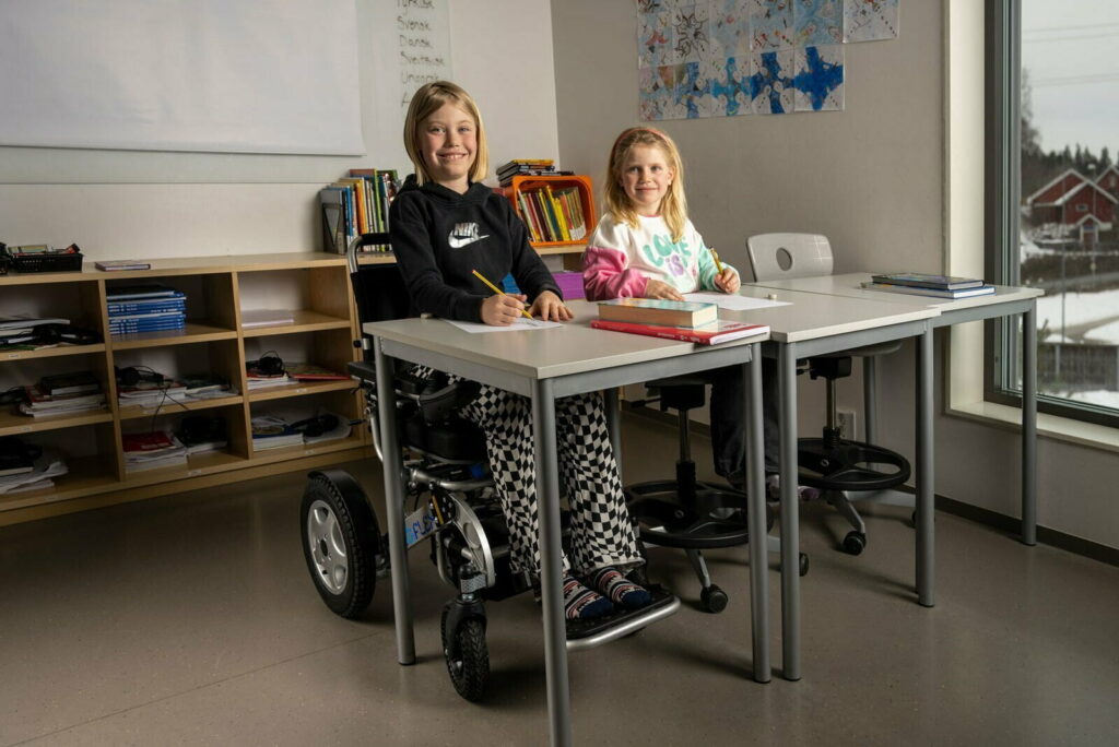 da och Sofia i skolan, med Ida som ler mot kameran medan hon sitter i sin Eloflex elrullstol vid sina skolbänkar. Bakom dem står en låg bokhylla med skolböcker, och på väggarna hänger färgglada teckningar.