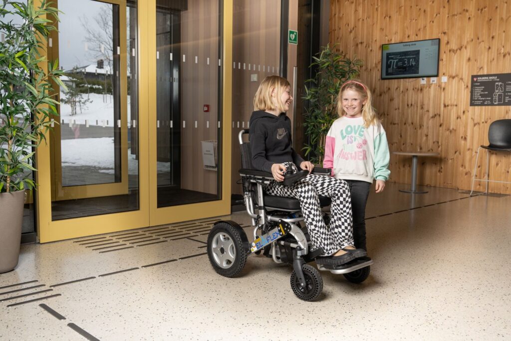 Ida ser på Sofia. Ida smiler til lillesøsteren, mens Sofia ser mot kameraet. Ida sitter i en eloflex rullestol. De står akkurat innenfor inngangsdøra til skolen.