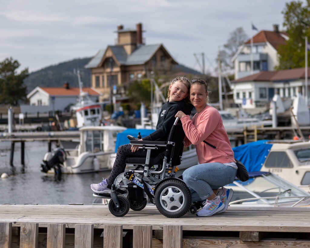 Heidi och Ida ler mot kameran medan de sitter vid en brygga. Ida sitter i en rullstol med hus och båtar i bakgrunden